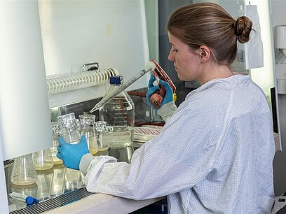 Eine junge Frau steht im Labor mit einer großen Pipette und einem Erlenmeyerkolben in den Händen.