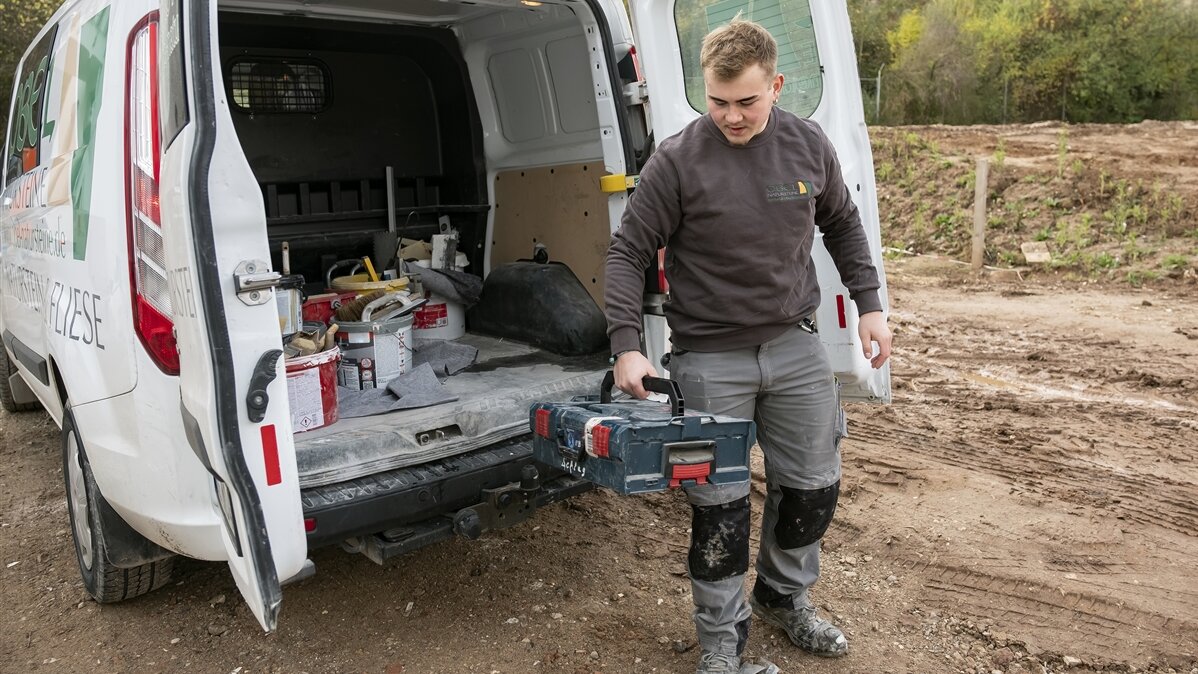 Clemens trägt in Arbeitskleidung einen Werkzeugkasten vom Auto zur Baustelle.