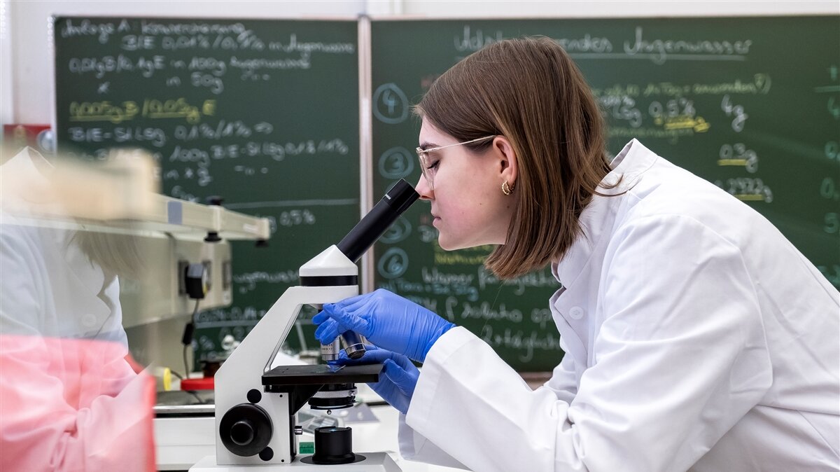 Eine junge Frau im Laborkittel untersucht eine Probe unter dem Mikroskop. 