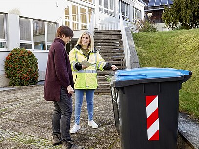 Eine Frau in einer Warnjacke erklärt einer anderen Frau die Mülltrennung. 