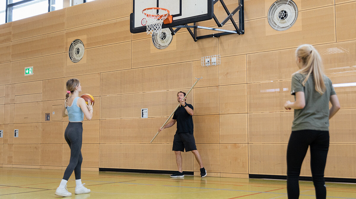Ein junger Mann verstellt mit einer Stange die Höhe eines Basketballkorbs in einer Turnhalle.