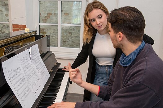 Eine junge Frau gibt einem jungen Mann Klavierunterricht.