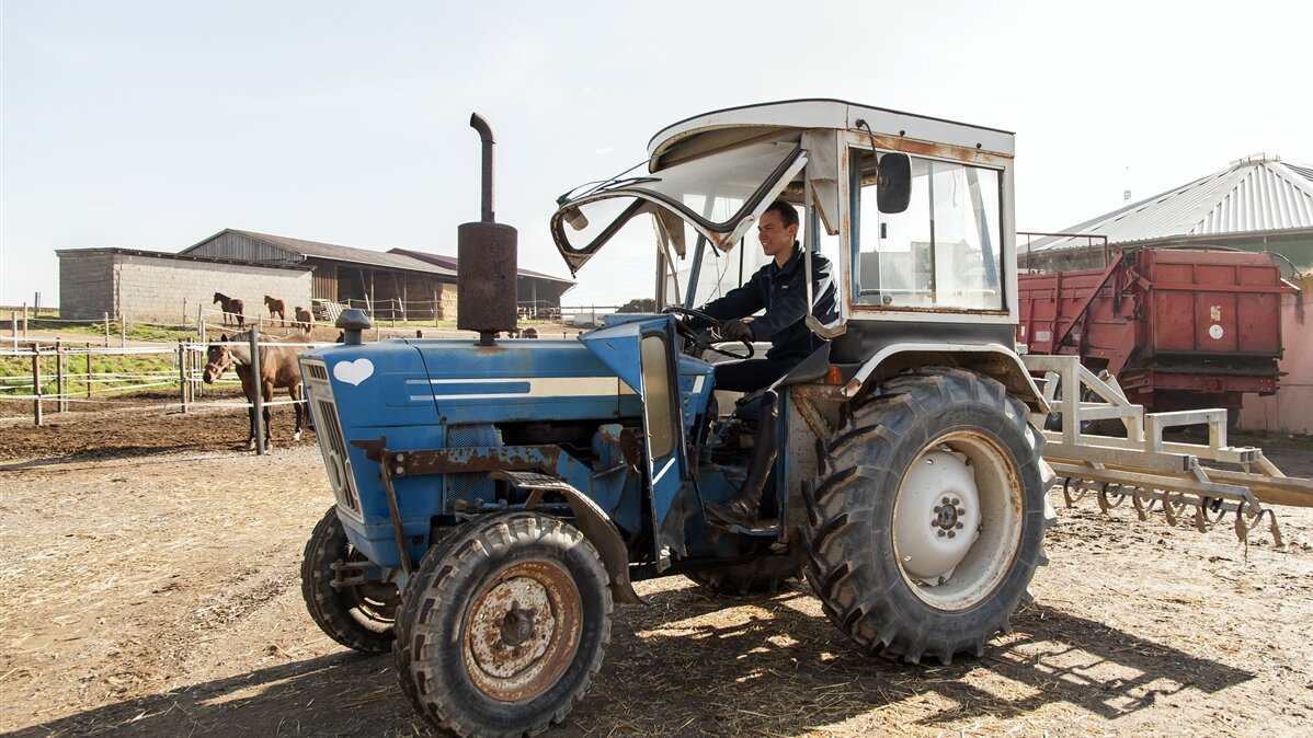 Ein Landwirt fährt mit dem Traktor auf einem Hof an einer Pferdekoppel entlang.