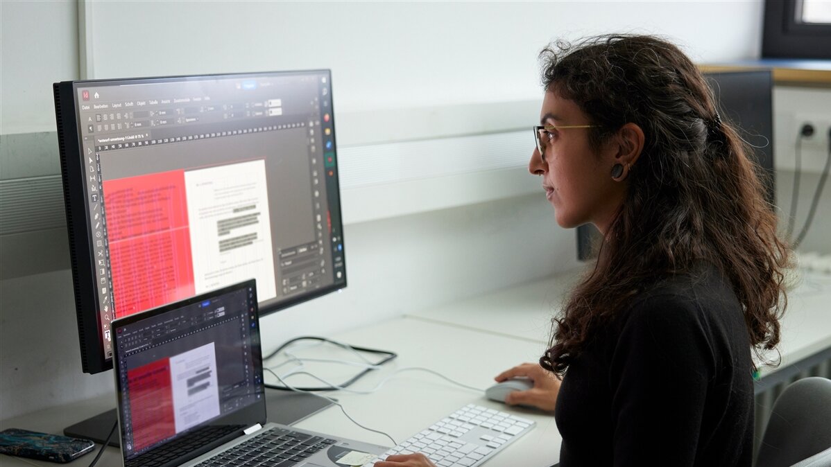 Yasmin sitzt vor zwei Computerbildschirmen und gestaltet das Programmheft.