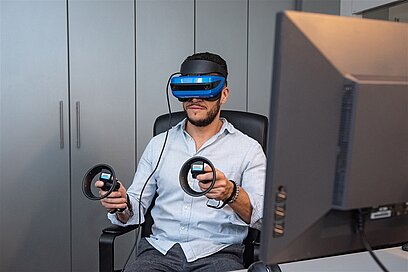 Ein Mann trägt im Sitzen eine VR-Brille und hält in jeder Hand einen Controller.
