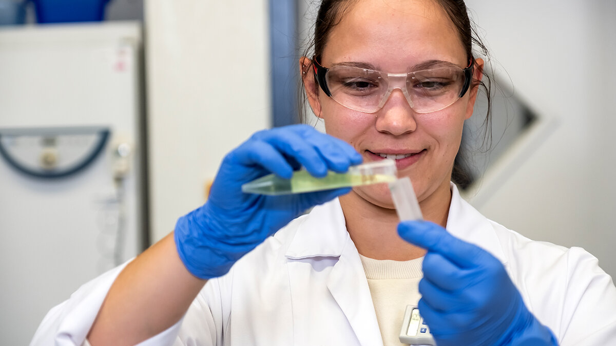 Eine junge Frau mit Schutzbrille füllt im Labor Flüssigkeit von einem Reagenzglas in ein anderes.