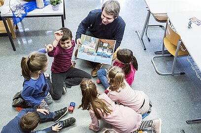 Ein Mann betreut eine Gruppe Kinder und liest ihnen  etwas vor.
