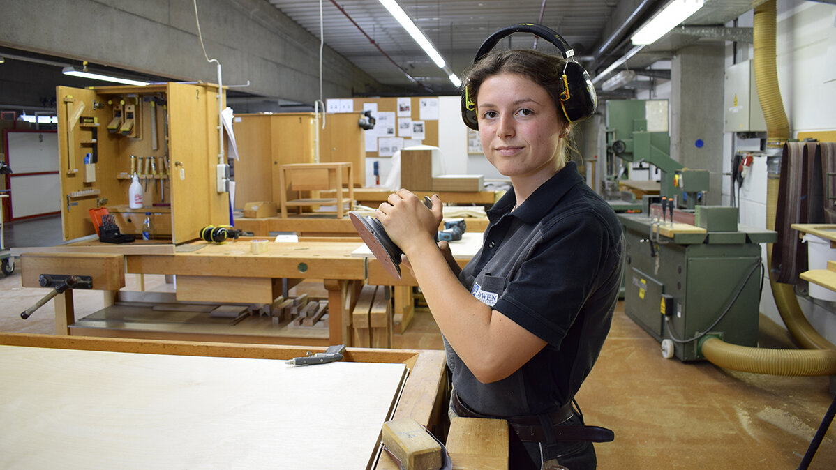 Judika steht  mit Gehörschutz in der Werkstatt und hält ein Schleifgerät in der Hand.