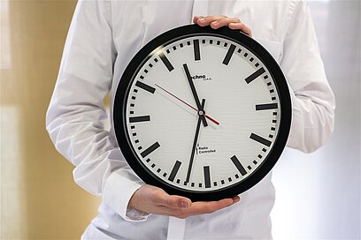 Eine Person hält eine große Uhr in den Händen, es ist halb Zwölf.