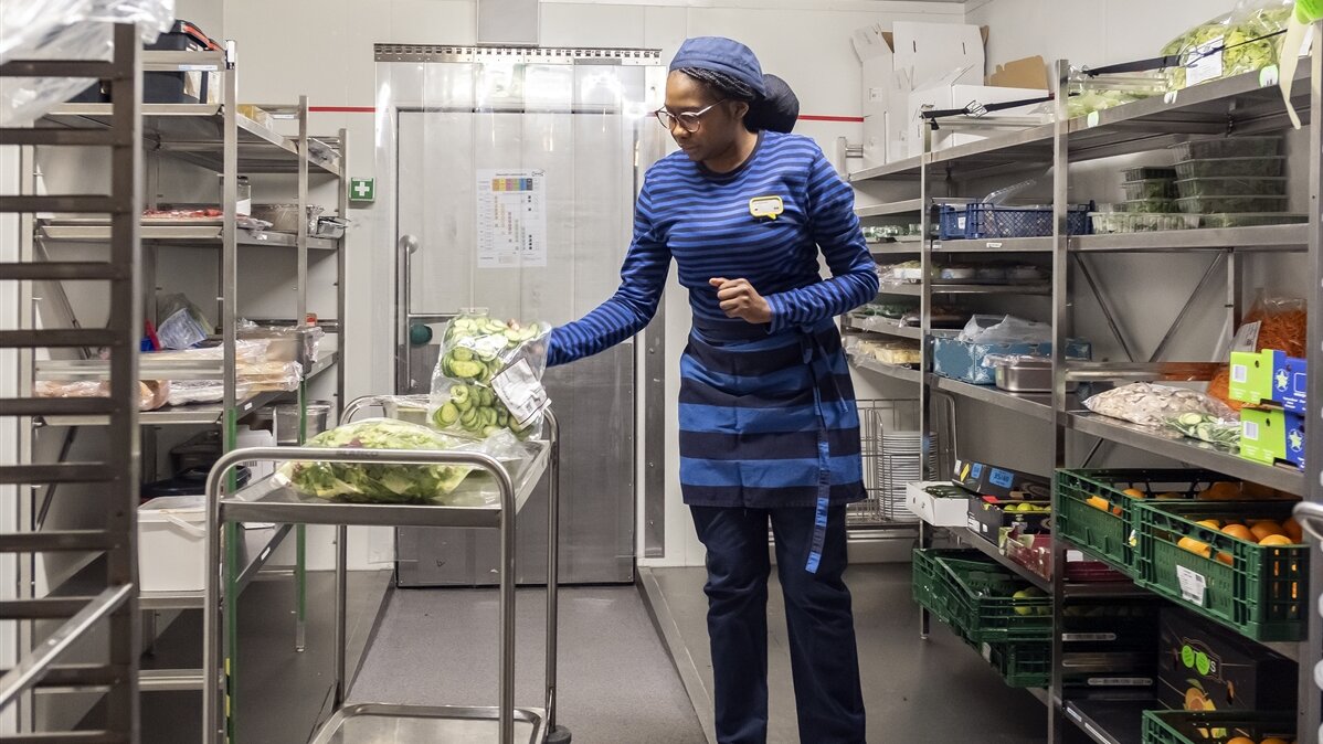 Josephat legt in Arbeitskleidung im Kühlraum Salatzutaten auf einen Metallwagen.