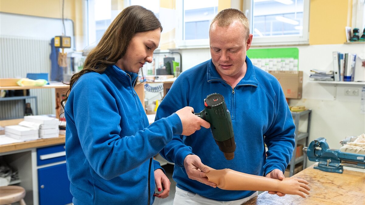 Ein Mann und eine Frau in einer Werkstatt bringen mit einem Heißluftföhn einen Kosmetikhandschuh an einer Handprothese an.