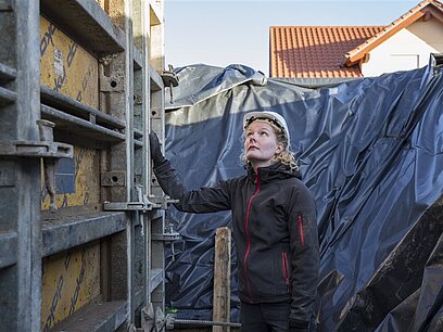 Eine junge Frau mit Schutzhelm prüft die Metallspannung einer Außenwand auf einer Baustelle.