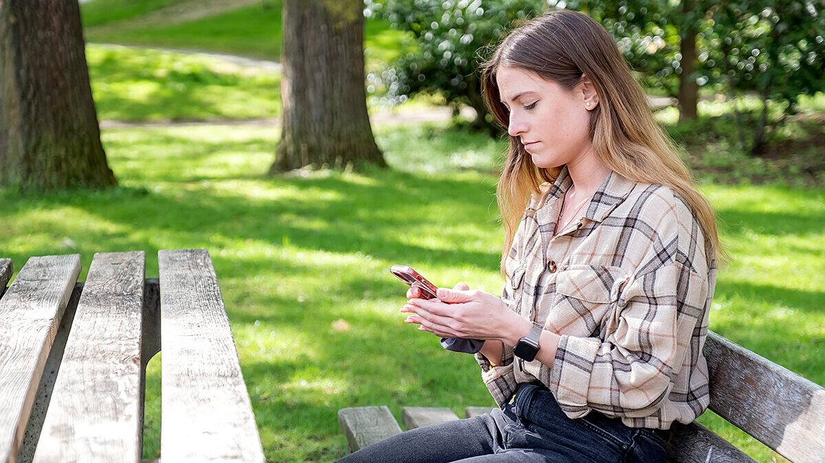 Eine junge Frau sitzt auf einer Bank und schaut auf ein Handy.