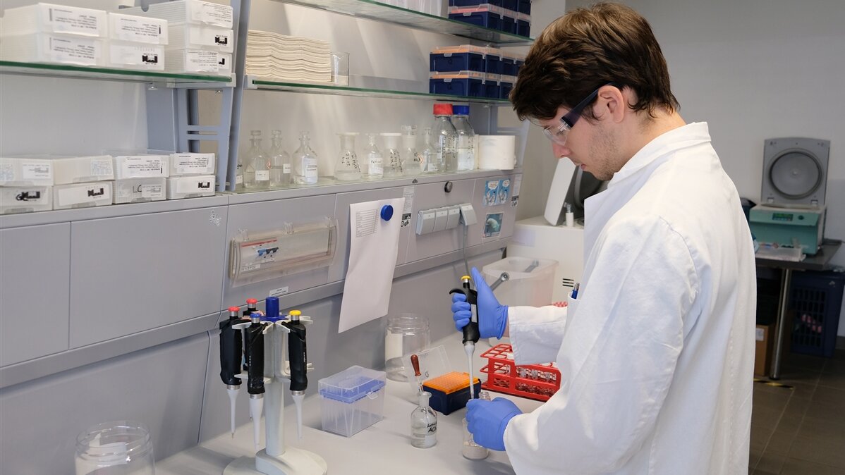 Ein junger Mann im Laborkittel füllt im Labor mit einer Pipette eine Flüssigkeit in ein Glas.