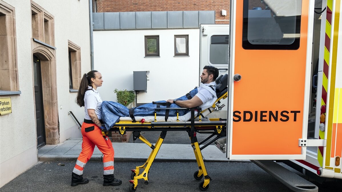 Eine junge Frau schiebt eine Trage mit einem verletzten Mann in einen Krankenwagen.