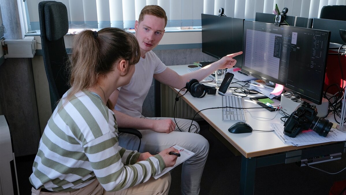 Ein junger Mediengestalter zeigt seiner Kollegin eine Aufgabe an seinem Computer.