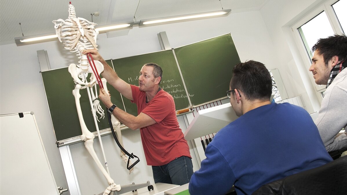 Ein Lehrer zeigt zwei Schülern etwas am menschlichen Skelett.