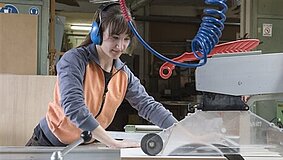 Eine junge Tischlerin mit Ohrschützern bearbeitet eine Holzplatte an einer Maschine.