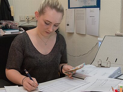 Eine junge Frau sitzt an einem Schreibtisch, prüft Dokumente und macht Notizen.