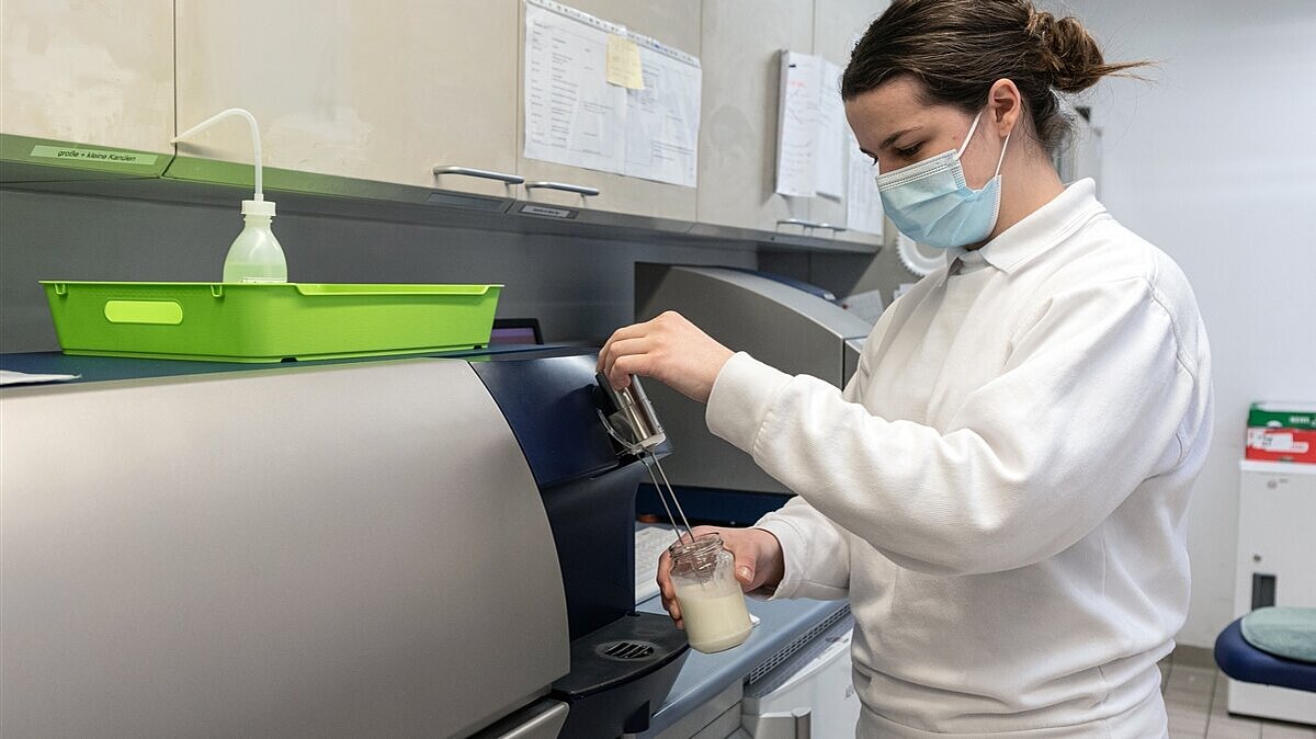 Eine junge Frau untersucht mit Atemschutzmaske und einem Milkosan-Analysegerät eine Milchprobe.