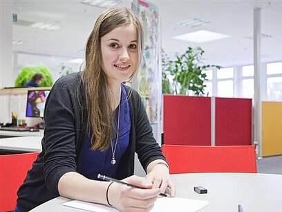 Eine junge Frau sitzt am Tisch vor einem Blatt Papier und hält einen Bleistift in der Hand.