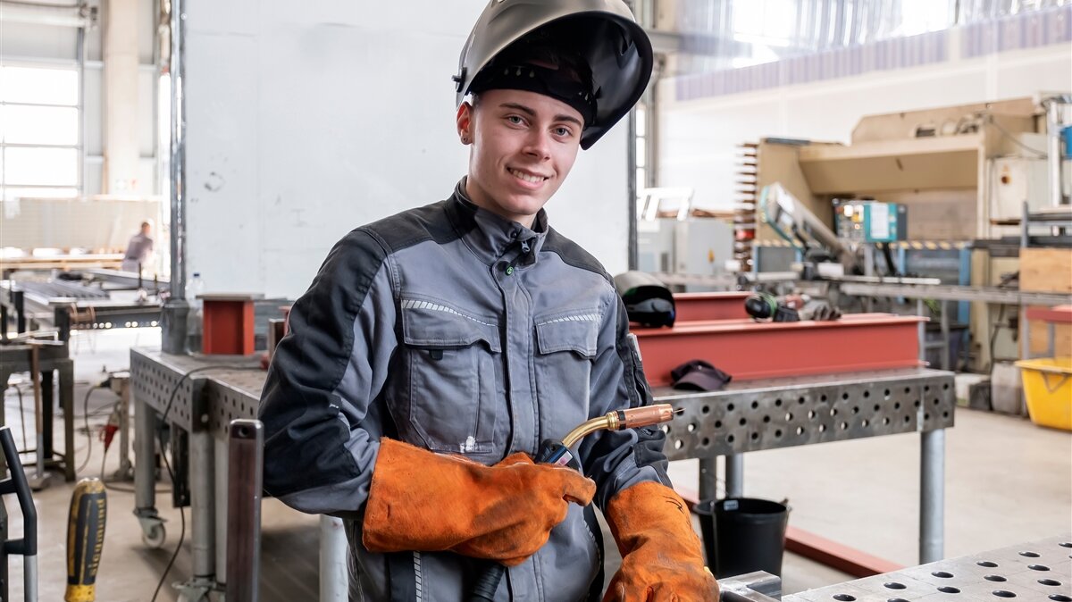 Ein junger Mann in Hitzeschutzkleidung hat ein Schweißgerät in der Hand und lächelt in die Kamera.