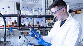 Eine Fachkraft Abwassertechnik mit Handschuhen prüft im Labor eine Flüssigkeit in einem Reagenzglas.