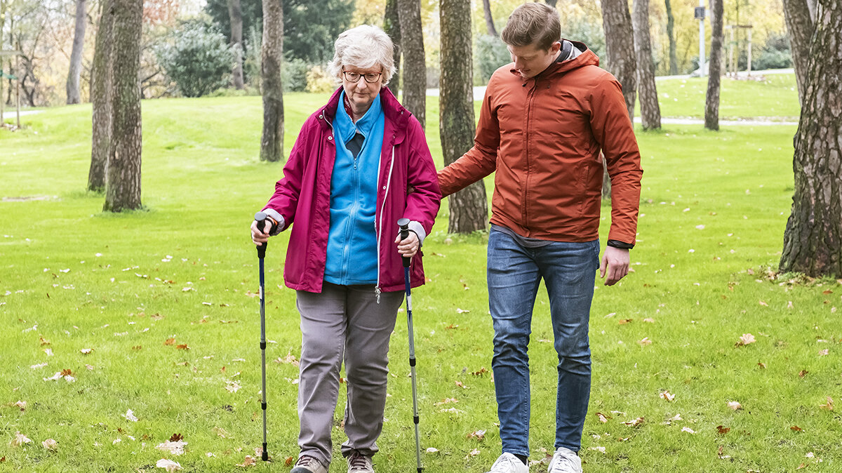 Ein junger Mann unterstützt eine ältere Frau bei einem Spaziergang.
