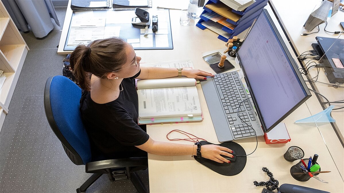 Eine junge Frau sitzt am Schreibtisch auf dem eine Akte liegt und arbeitet am Computer.