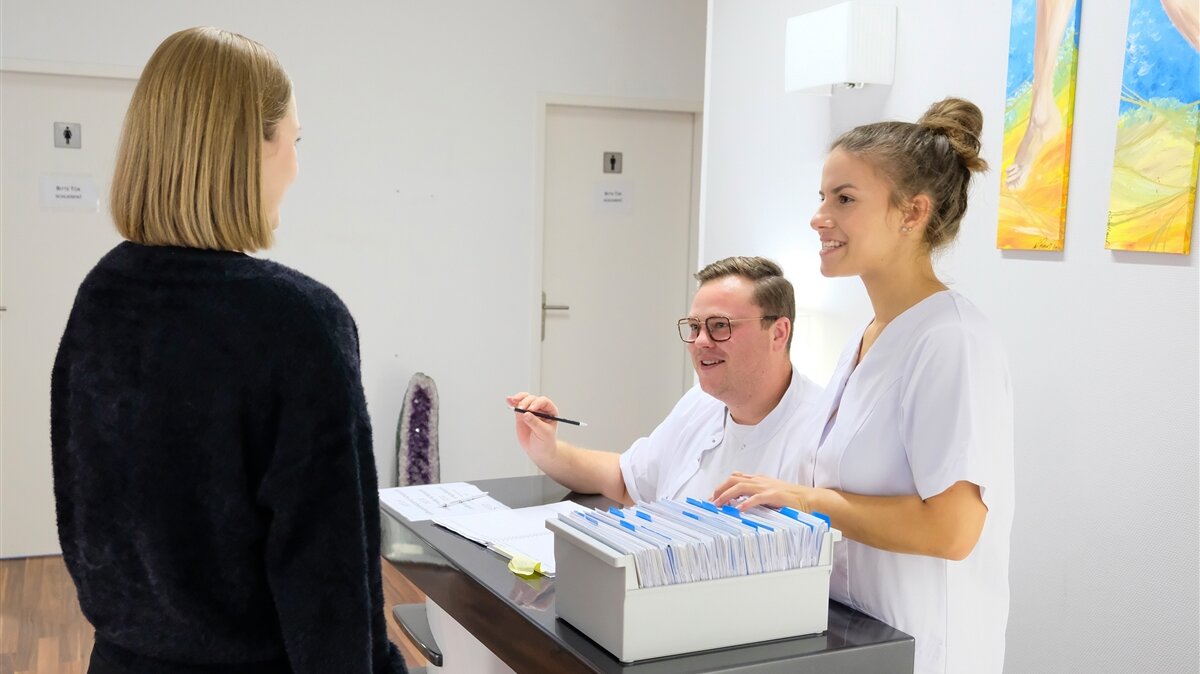 Marie-Louise sucht an der Anmeldung Patientenkarten, während ein Kollege eine Patientin annimmt.