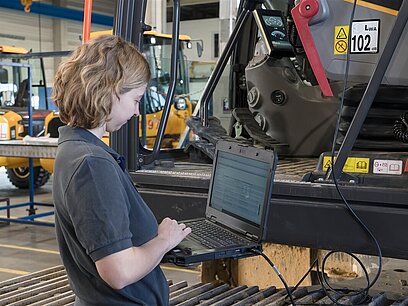 Eine junge Frau in einer Werkhalle liest mithilfe eines Laptops das System eines Fahrzeugs aus.