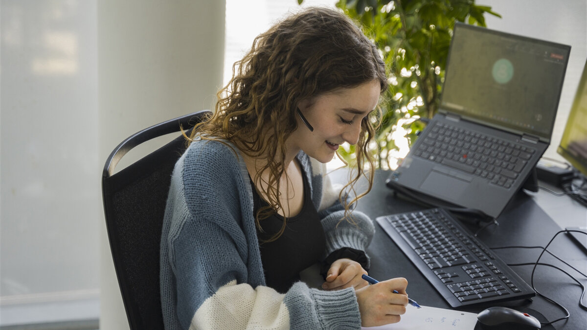 Eine junge Frau sitzt mit Headset in einem Online-Meeting am Schreibtisch und notiert sich etwas.