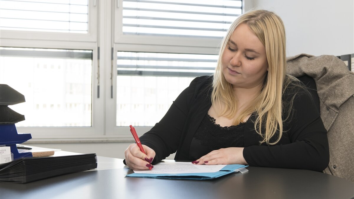 Eine Frau sitzt an einem Tisch und schreibt mit einem Stift auf ein Blatt Papier.