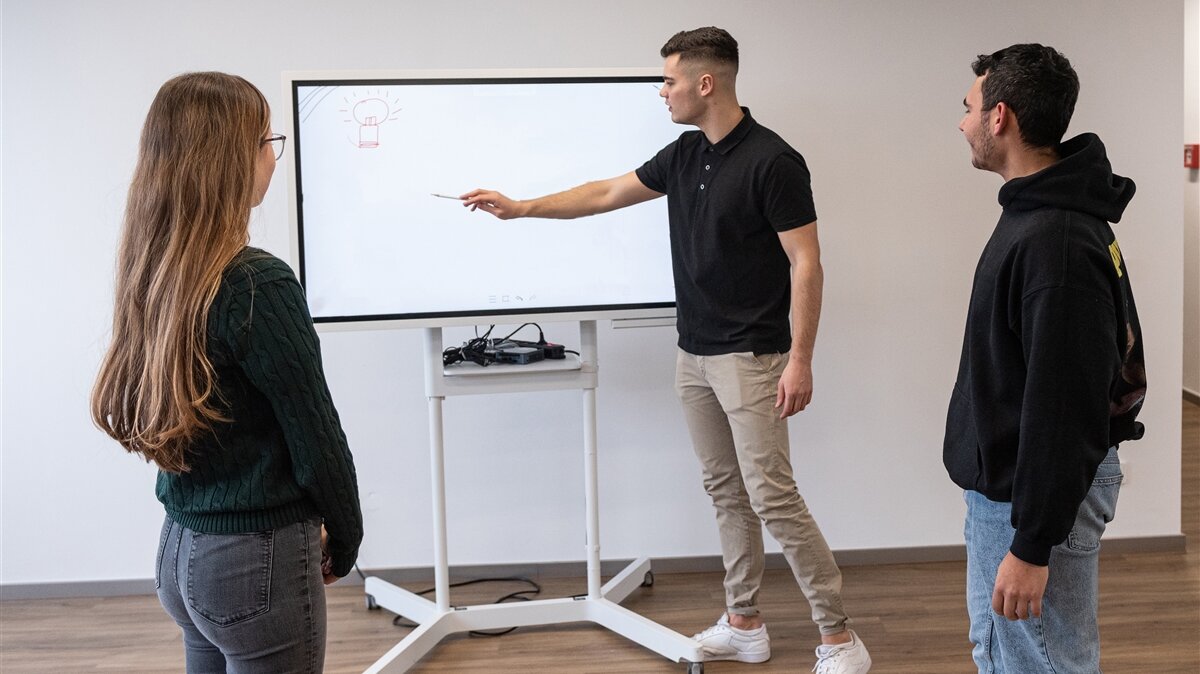 Ein junger Mann erklärt einer jungen Frau und einem Mann etwas auf einem Smartboard.