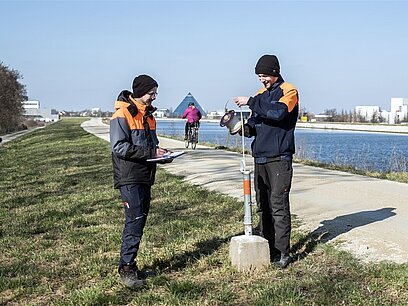 Zwei junge Männer tragen Schutzkleidung und stehen auf einem Weg an einem Kanal.