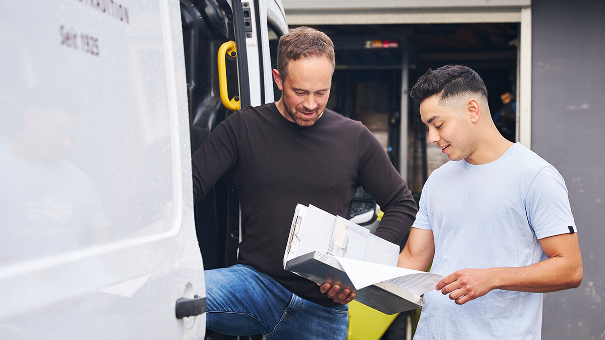 Ein jüngerer und ein älterer Mann lesen neben einem Transporter ein Auftragsbuch.