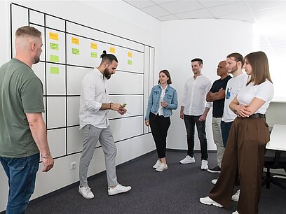 Ein junger Mann verteilt Postits an im Halbkreis vor ihm stehende junge Leute.