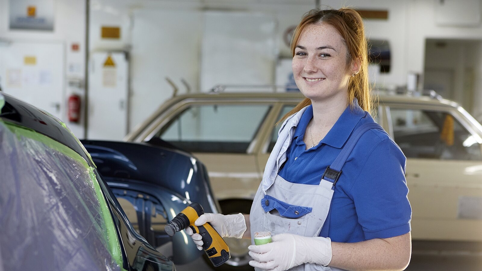 Eine Frau in Arbeitsklamotten hält ein Werkzeug in der Hand und lächelt in die Kamera.