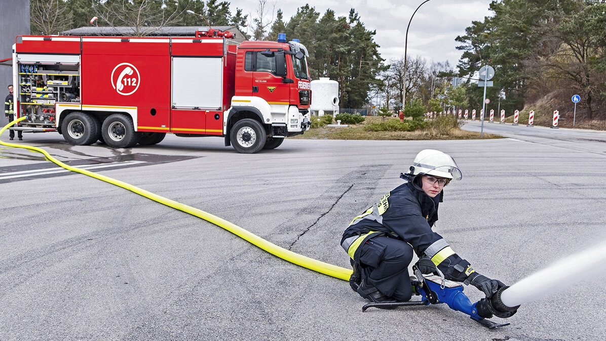 Eine Feuerwehrfrau in Schutzkleidung und Helm bedient den Schlauch eines Löschfahrzeugs.