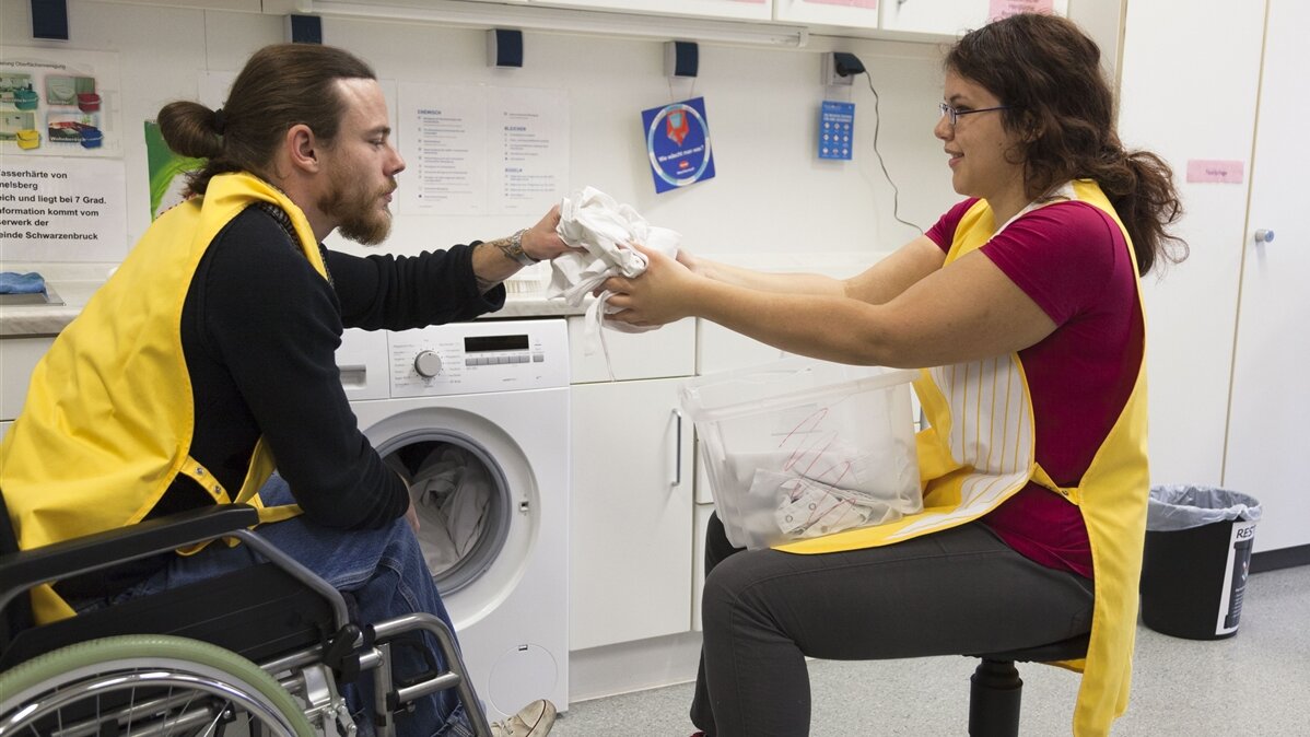 Eine junge Frau hilft einem Mann im Rollstuhl beim Wäsche waschen.