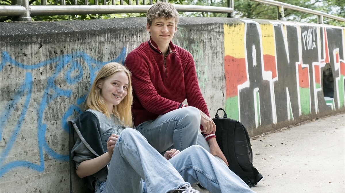 Zwei Jugendliche sitzen entspannt vor einer bunt besprühten Betonwand mit Graffiti. Beide lächeln in die Kamera.