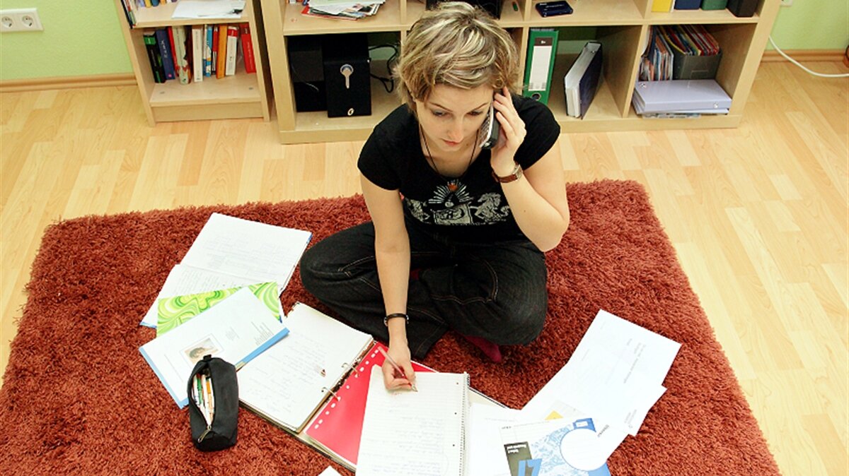 Eine junge Frau sitzt auf dem Boden, umgeben von Unterlagen, und telefoniert.