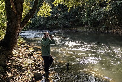 Ein junger Mann blickt mit einem Fernglas auf einen Fluss.