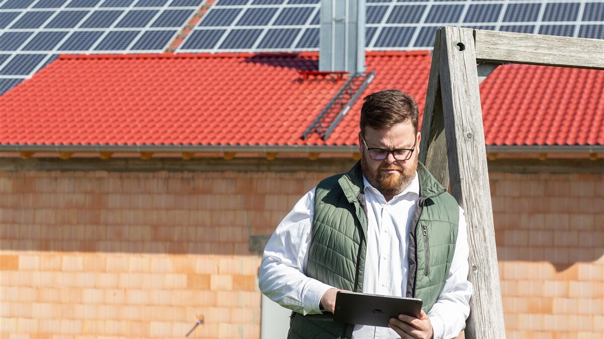 Ein Mann steht vor einem Haus mit Solar-Panels und arbeitet am Tablet.