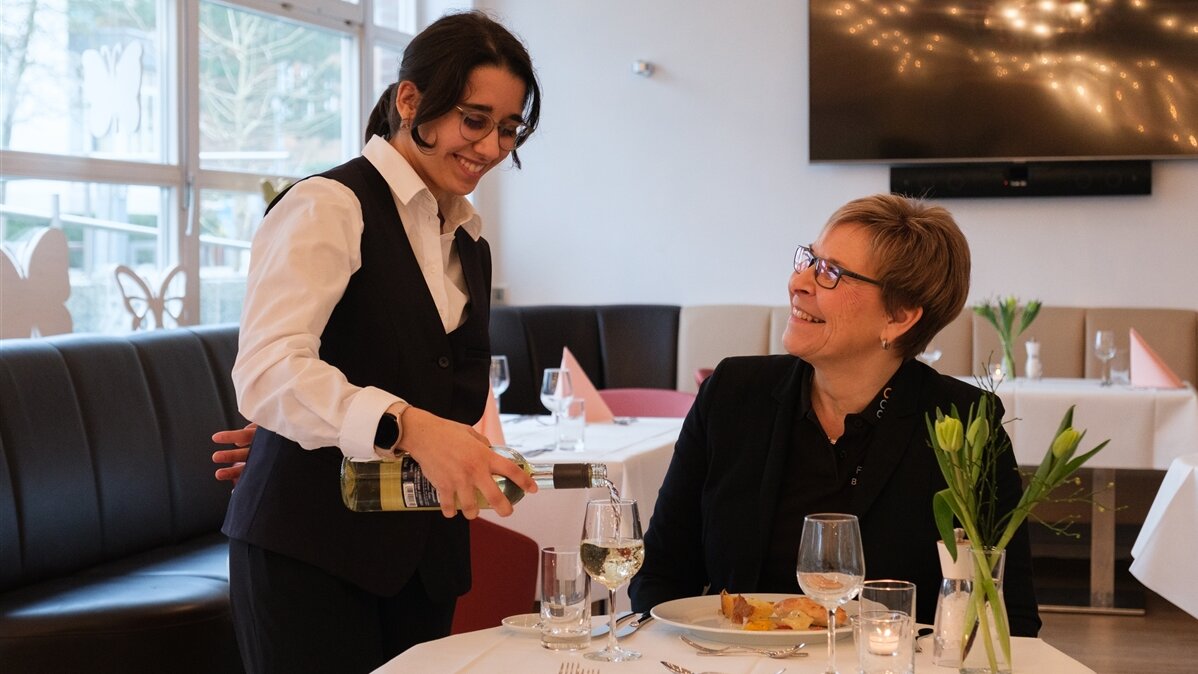 Eine junge Frau lächelt und schenkt einem Gast im Restaurant ein Glas Wein ein.