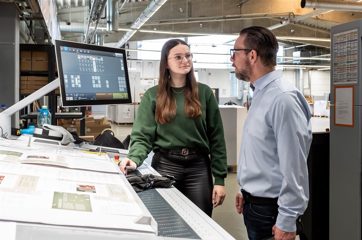 Eine junge Frau und ein Mann stehen vor einer Offsetdruckmaschine und sprechen miteinander.