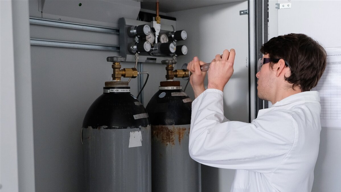 Ein Mann in Laborkittel arbeitet mit einem Schraubenschlüssel an einer großen Gasflasche.