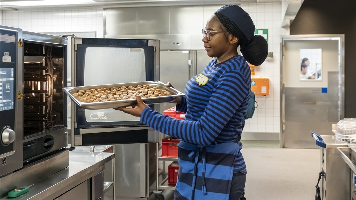 Josephat trägt Arbeitskleidung und schiebt ein Tablett mit Fleischbällchen in den Ofen.