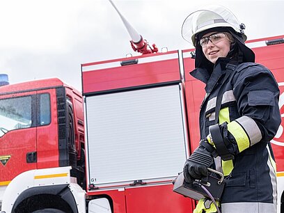 Eine Frau in Feuwerwehrausrüstung steht vor einem Feuerwehrfahrzeug.