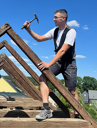 Ein junger Mann arbeitet mit einem Hammer auf einem Dachstuhl.
